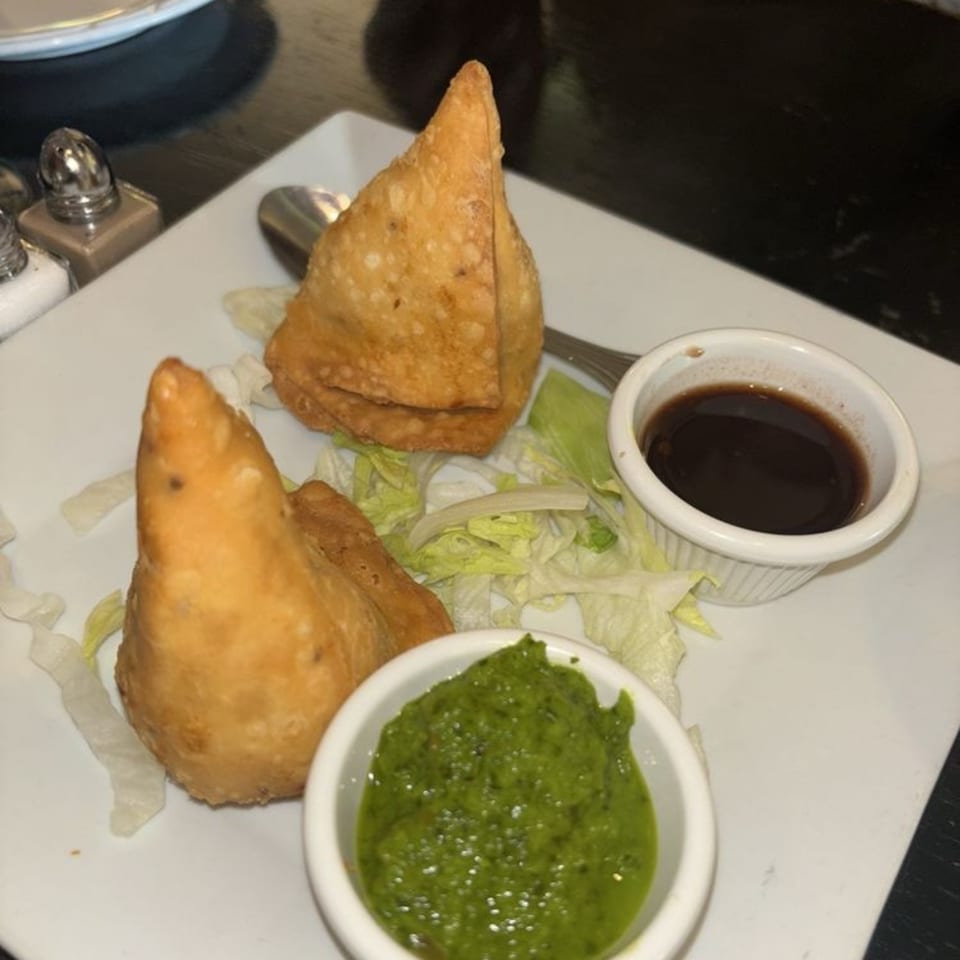 Two golden-brown samosas with green chutney and tamarind sauce on a white plate with lettuce.