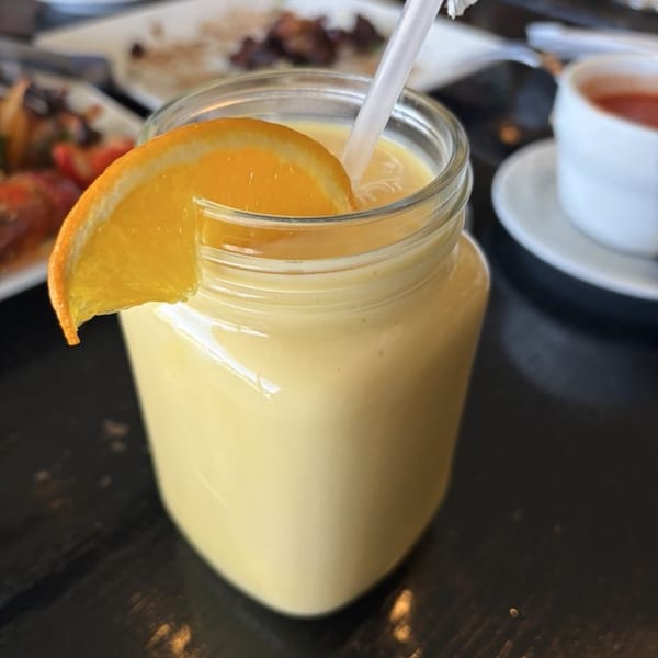 A glass jar of mango lassi with an orange slice and straw, on a dark table.