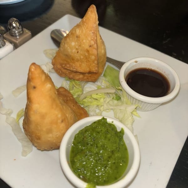 Two golden-brown Indian samosas with green and dark dipping sauces on a white plate.